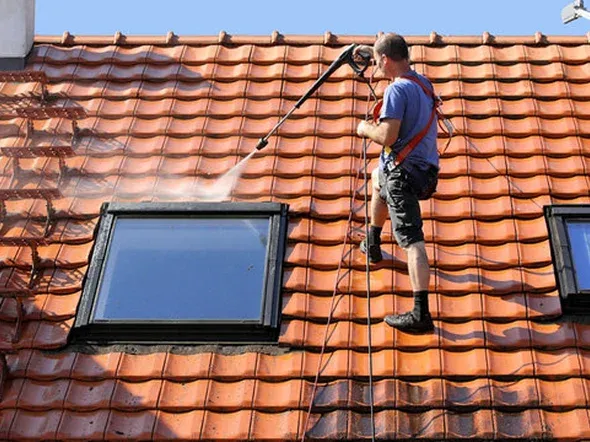 Man pressure washing a tiled roof near a window